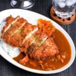Japanese chicken katsu curry served on a white oval-shaped plate with beige rim, next to a wooden spoon on a black background topdown view