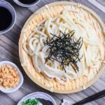 Cold udon noodles on a bamboo zaru (tray) next to homemade mentsuyu dipping sauce, tenkasu (tempura bits), green onion, wasabi and grated ginger