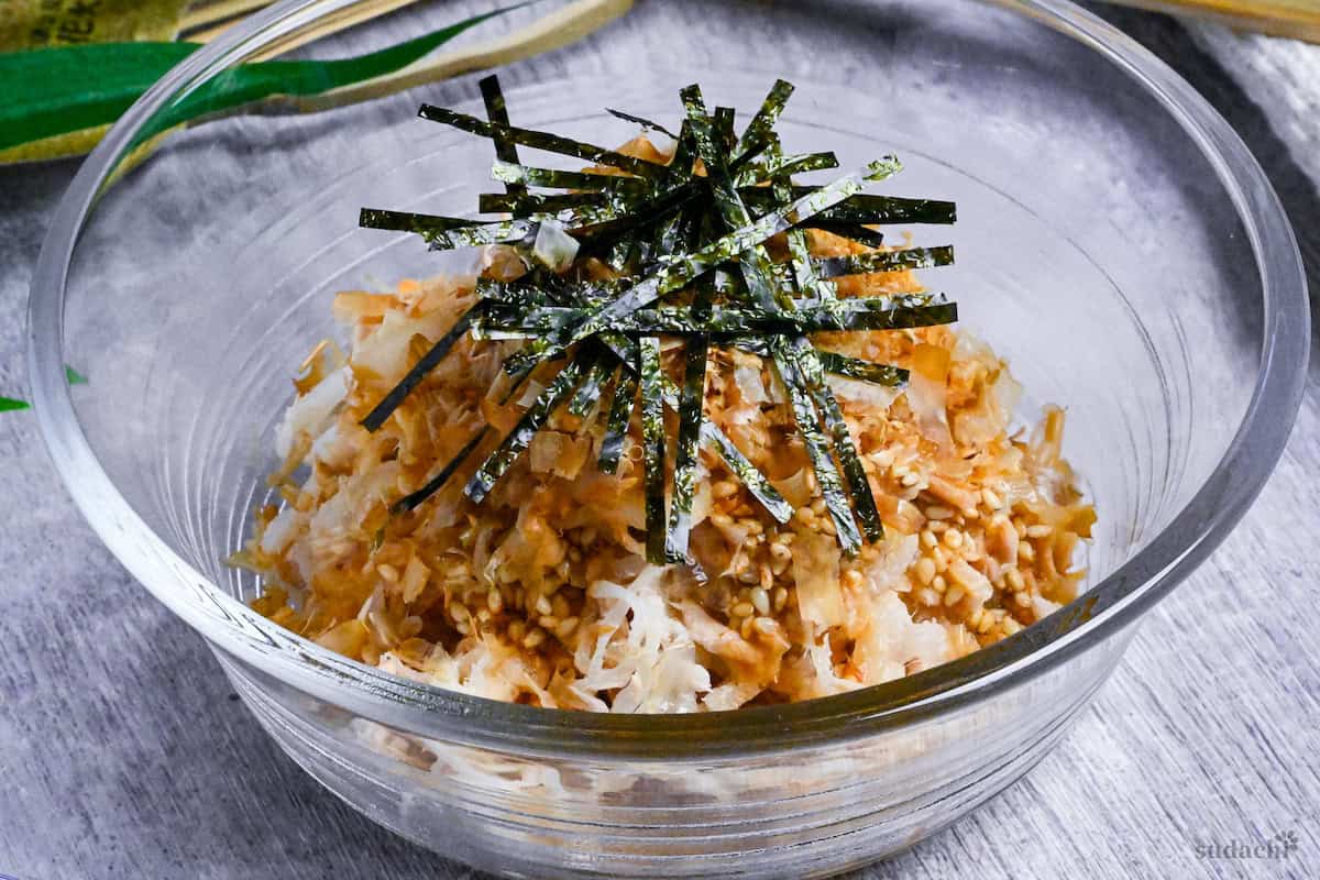 Addictive daikon radish salad topped with bonito flakes, sesame seeds and kizami nori served in a glass bowl next to black chopsticks and a Japanese fan in the background