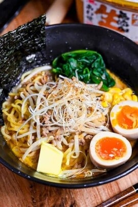 Easy homemade pork miso ramen in a black bowl topped with ramen eggs, spinach, beansprouts, nori and sweetcorn next to a plate of fried gyoza and a tub of miso paste.