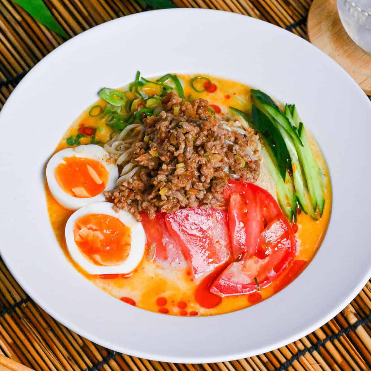 Hiyashi tantanmen in a white dish topped with boiled egg halves, julienned cucumber, chopped green onion, sliced tomato and seasoned ground pork on a bamboo mat background next to a glass of iced water and light wooden chopsticks
