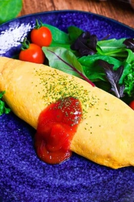Omurice (Japanese omelette rice) topped with ketchup and served on a blue plate next to salad leaves, baby tomatoes and fresh parsley