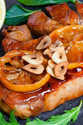 Lemon Teriyaki Pork Chop topped with lemon slices and fried garlic chips on a black slate-style square plate with pan fried mushrooms and salad leaves