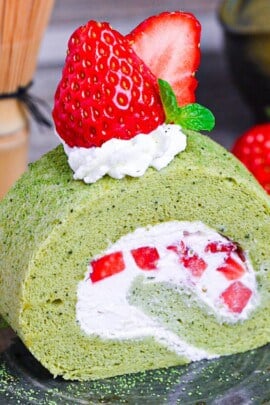 A slice of Matcha Roll Cake (Green Tea Swiss Roll) on a green plate topped with cream, strawberries and a mint leaf