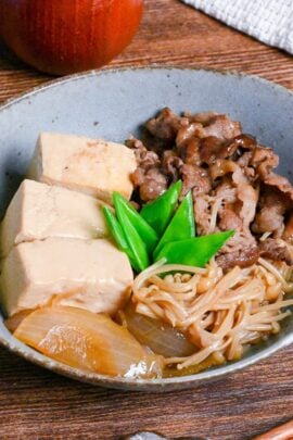 niku dofu in a eggshell blue bowl made with firm tofu, thinly sliced beef, onions and enoki mushrooms simmered in a sweet and salty Japanese broth and topped with blanched snow peas on a wooden table