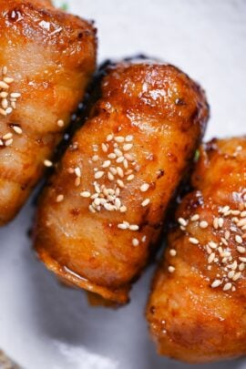 Three nikumaki onigiri (pork belly wrapped rice balls) arranged on a cream plate decorated with Perilla leaves and sprinkled with sesame seeds