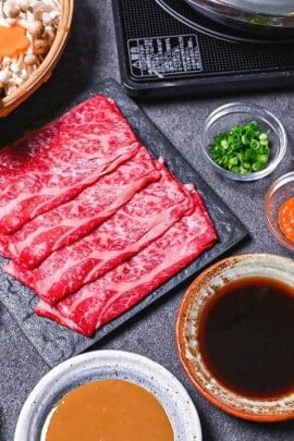 Ingredients to make shabu shabu on a gray background (a basket of vegetables and noodles, a plate of thinly sliced wagyu beef, various bowls of condiments and a bowl of sesame sauce and ponzu sauce)