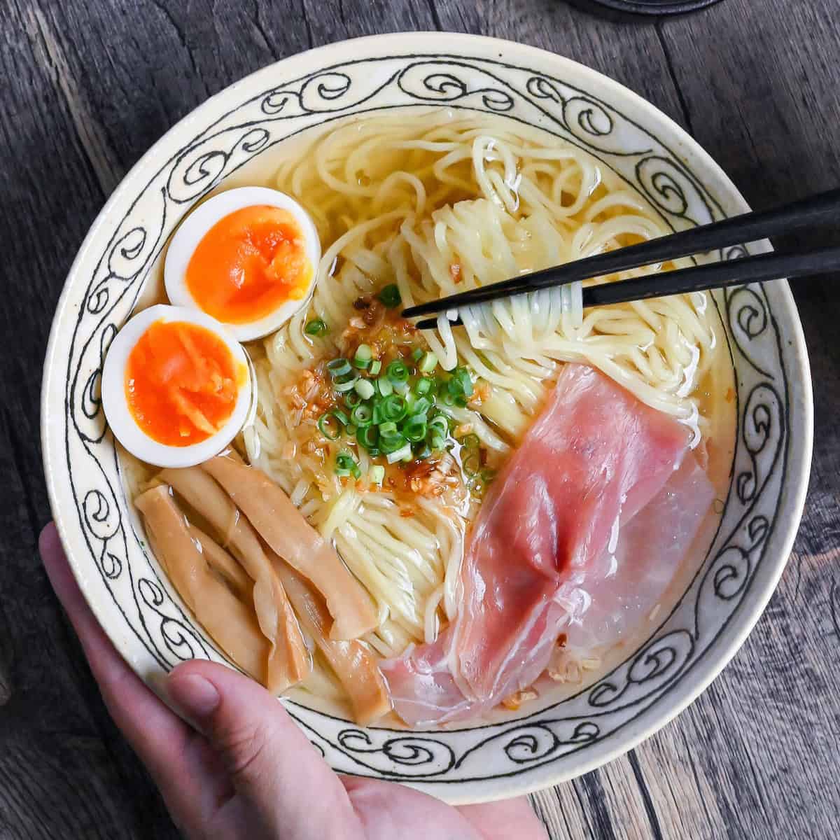 Homemade shio ramen topped with soft boiled eggs, menma, and ham in a cream bowl with black patterned rim on ashy wood background