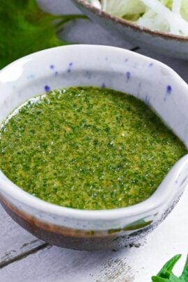 Japanese shiso dressing in a small blue and brown ceramic jug with a bowl of salad in the background and shiso leaves scattered around