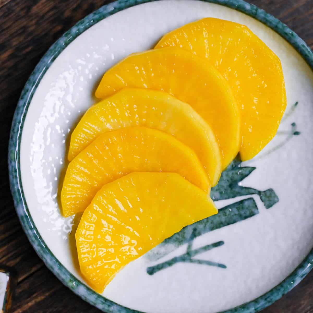5 pieces of yellow pickled daikon radish (takuan) cut into half moons and arranged in a row on a white plate with green ink design close up