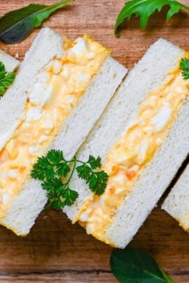 Four Japanese egg finger sandwiches (tamago sando) on a wooden chopping board with salad leaves