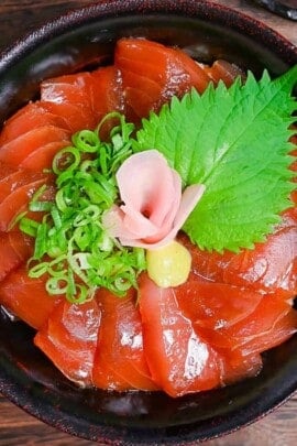 Tekkadon - vinegared rice topped with slices of fresh tuna sashimi, pickled ginger, wasabi, shiso and chopped spring onion in a black lacquerware bowl