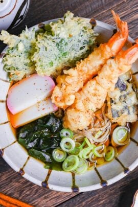 Tempura soba topped with tempura shrimp and vegetables, chopped green onions, kamaboko fishcakes and wakame seadweed in a striped bowl on a dark wooden effect background top down view