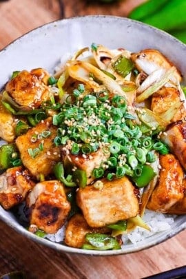 tofu donburi with teriyaki glaze topped with chili oil, green onion and sesame seeds over rice in a mottled bowl