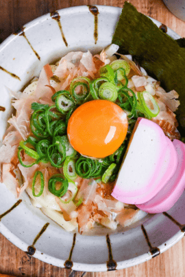 abura udon topped with bonito flakes, chopped green onions, nori, kamaboko fishcakes and a raw egg yolk served in an off-white bowl with brown stripes top down thumbnail