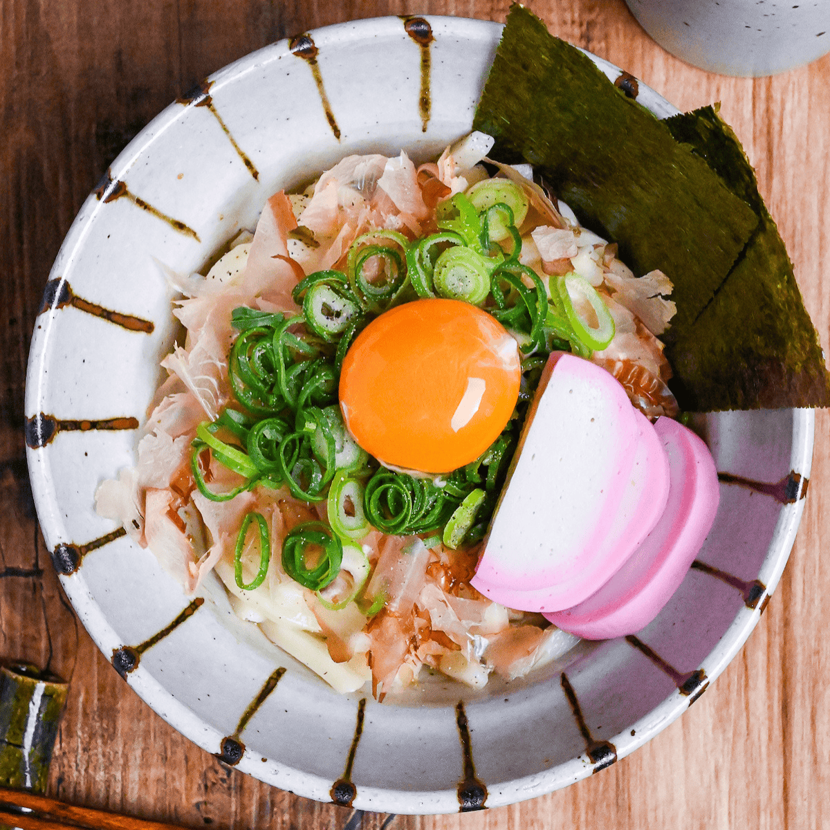 abura udon topped with bonito flakes, chopped green onions, nori, kamaboko fishcakes and a raw egg yolk served in an off-white bowl with brown stripes top down thumbnail