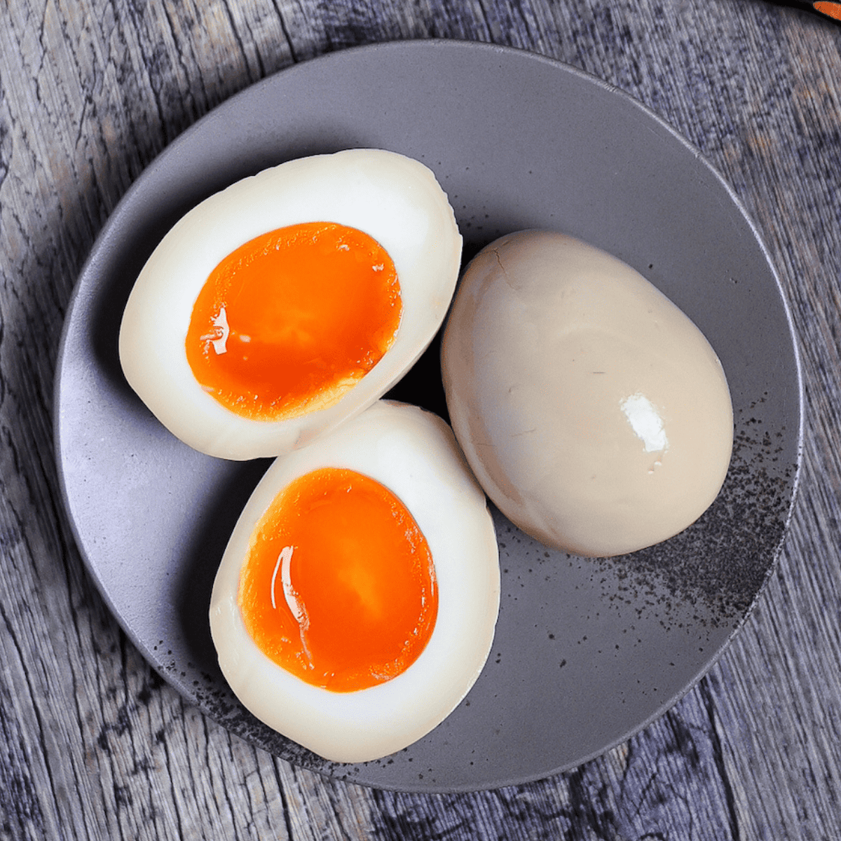 Two Ramen Eggs (Ajitsuke Tamago) with one cut in half to reveal jelly yolk on a gray plate next to wooden chopsticks on a ash-wood background