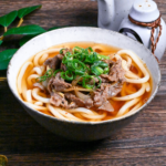 Beef niku udon topped with green onions in a white and gray mottled ceramic bowl on a dark wooden table side view