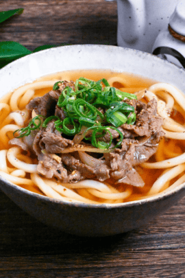 Beef niku udon topped with green onions in a white and gray mottled ceramic bowl on a dark wooden table side view