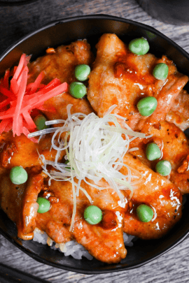 Japanese pork bowl (butadon) made with caramelized pork belly slices, green peas, pink pickled ginger and strings of Japanese leek in a black bowl on a wood-effect background