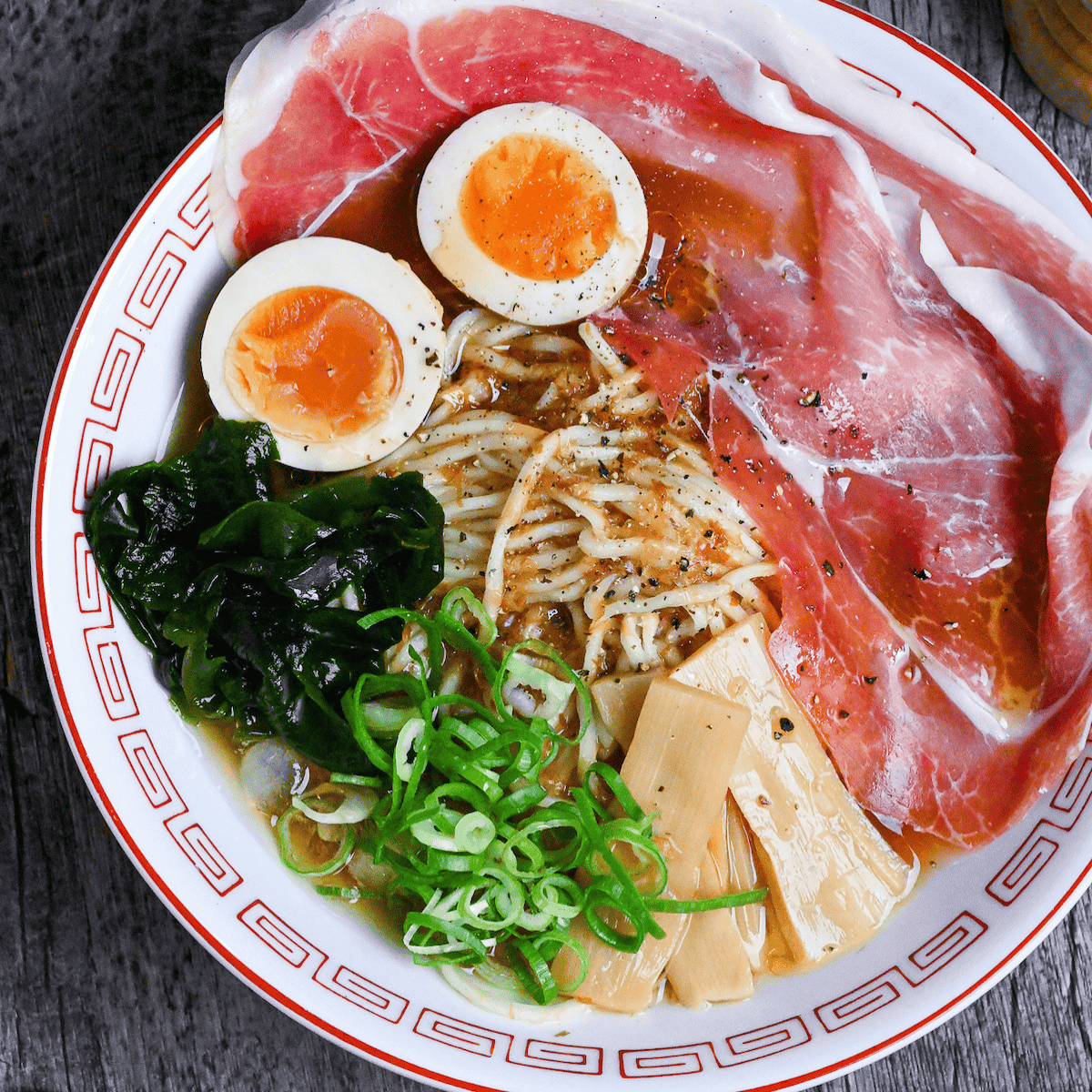 Cold ramen served in a white and red ramen bowl topped with cured ham, soft boiled eggs, wakame, chopped green onions and menma bamboo shoots on an ashy wood background