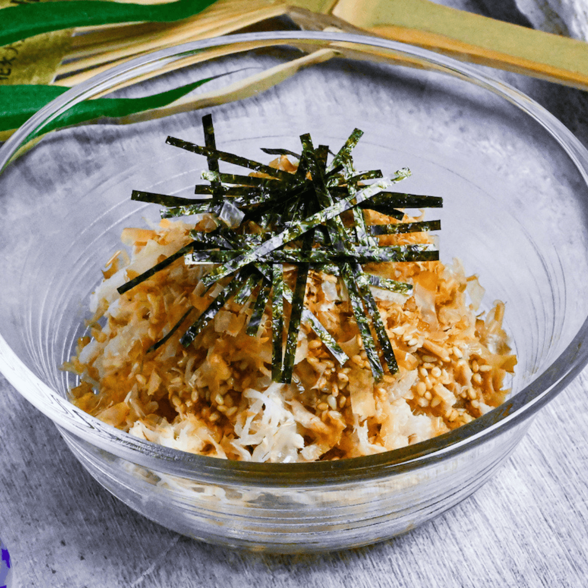 Addictive daikon radish salad topped with bonito flakes, sesame seeds and kizami nori served in a glass bowl next to black chopsticks and a Japanese fan in the background