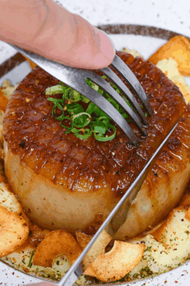 Cutting into a daikon steak served over mashed potato and sprinkled with crispy garlic chips on a white plate with gold rim