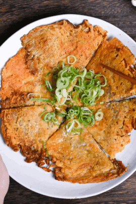 Crispy enoki cheese cut into triangles and served on a white plate with chopped green onions and a small bowl of ketchup on the side. One hand is holding the plate and the other is holding up one piece of enoki cheese with wooden chopsticks.