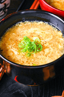 Kakitamajiru (Japanese egg drop soup) topped with chopped green onions and shichimi togarashi served in a black soup bowl on a black tray with wooden chopsticks