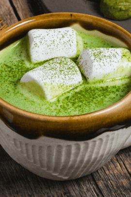 Matcha white hot chocolate in a brown and cream Japanese ceramic mug topped with marshmallows and matcha powder on a dark wooden background