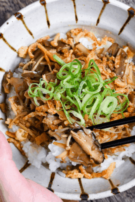 a hand holding mushroom donburi with black chopsticks in the other hand holding up mushrooms and rice