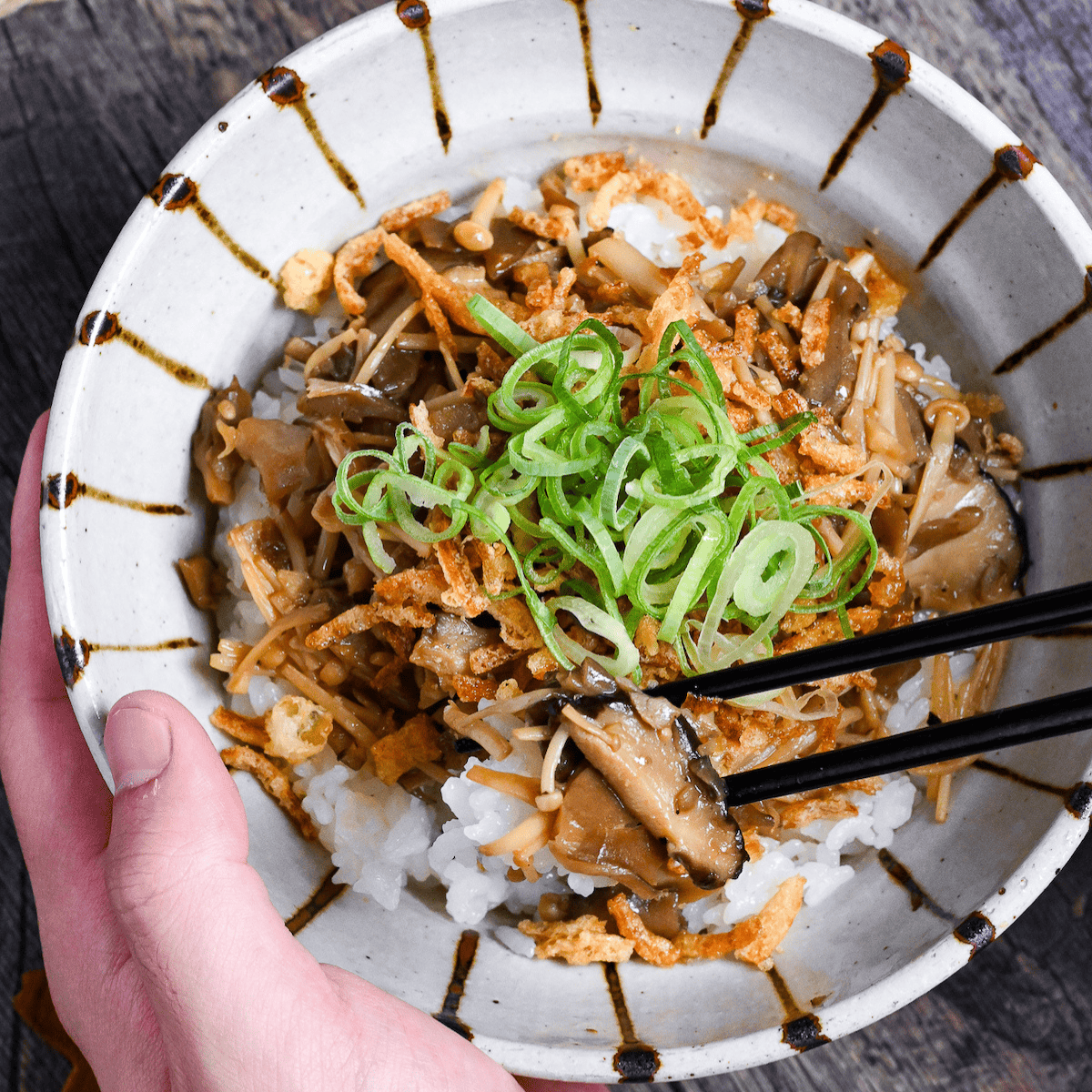 a hand holding mushroom donburi with black chopsticks in the other hand holding up mushrooms and rice