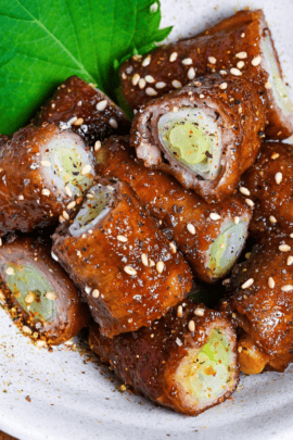 Glazed beef negimaki (beef leek rolls) cut into bitesize pieces in a white bowl decorated with a shiso leaf and sprinkled with shichimi togarashi and sesame seeds on a wooden background