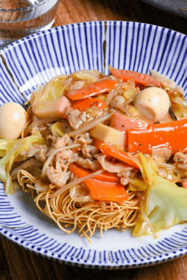 Sara udon served on a white and blue striped plate on a wooden surface next to black chopsticks and a glass of water