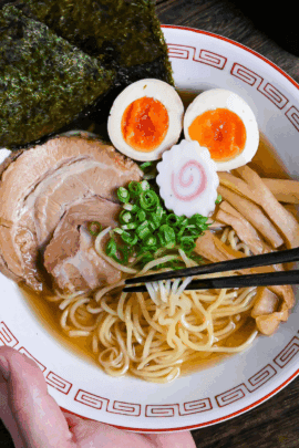 Homemade shoyu ramen in a white and red ramen bowl topped with pork chashu, ramen eggs, narutomaki, nori, chopped green onions and menma with noodles lifted with black chopsticks