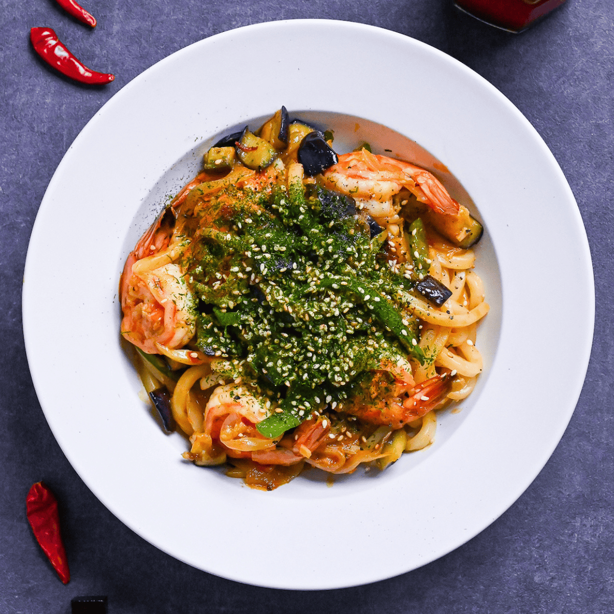 Spicy seafood yaki udon sprinkled with aonori and sesame seeds in a white dish on a gray surface surrounded by dry chilis and a jar of chili oil