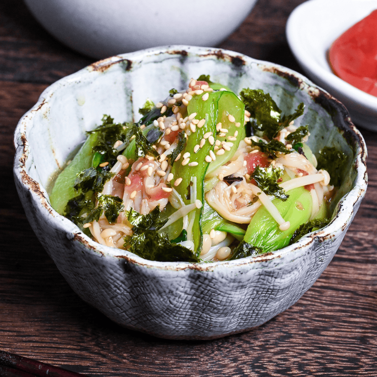 Wasabi-Ae salad with enoki mushrooms and bok choy in a blue and brown bowl on a dark wooden background with umeboshi on a white dish in the background